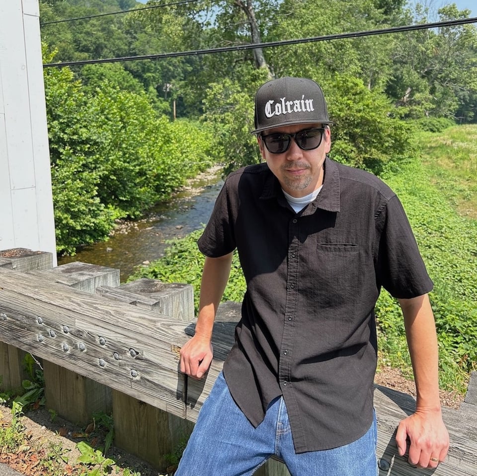 A man in blue jeans, an untucked black button-down shirt, sunglasses and a black baseball cap that says "COLRAIN' in white letters, leans against the railing of a bridge with a creek and woods behind him.