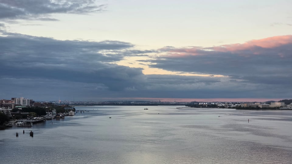 A wide river stretches toward a distant city skyline at dawn, its calm, silvery water dotted with small boats and channel markers, while a waterfront town with buildings and docks lines the left bank beneath a vast sky of layered grey clouds tinted pink and gold near the horizon.