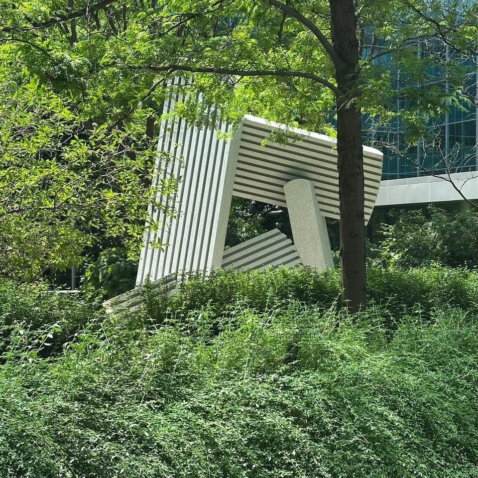 Photo of a sculpture consisting of lined metal blocks intersecting with each other, with lots of greenery surrounding them