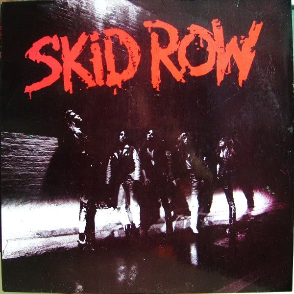 A black and white photo of the band on a sidewalk in front of a brick wall. The bottom of the wall, where it meets the sidewalk, is illuminated - the suggestion of car headlights perhaps? The ground is wet. The band members look in different directions.