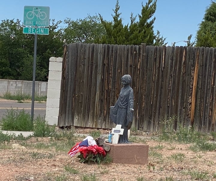 statue of young woman, white cross leaning on her legs. She sits on a concrete block. There are red flowers and American flag in the ground in front.