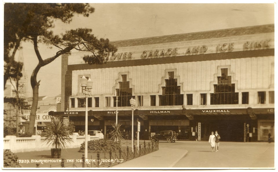 A huge hall. At the ground floor, a metal framed canopy advertises Morris, Hillman and Vauxhall cars. Above the walls have a wildly stylized ziggurat windows set into a concrete frame. Along the roof, neon letters spell out Westover Garage and Ice Rink.