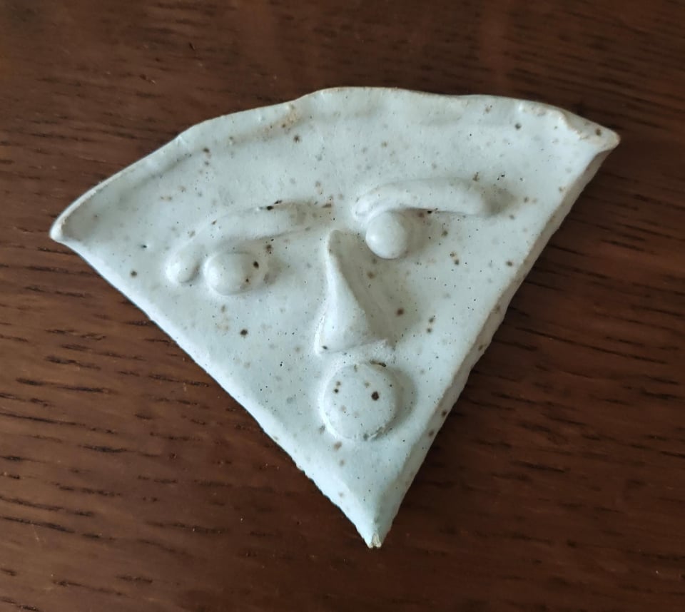 Close up photo of a wedge-shaped ceramics piece with eyes, eyebrows, nose and a round mouth attached. The whole piece is glazed off-white with brown speckles and it's photographed against a dark wood background