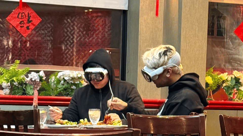 Two people eating lunch at a restuarant while both of them wear Vision Pro Headsets