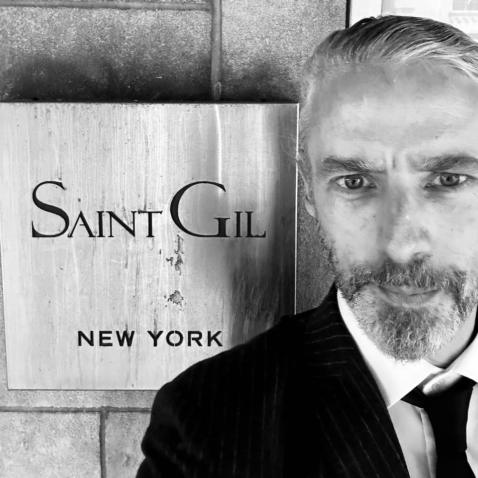 b/w photo of a white man in a suit & tie standing beside a sign that reads "Saint Gil - New York"