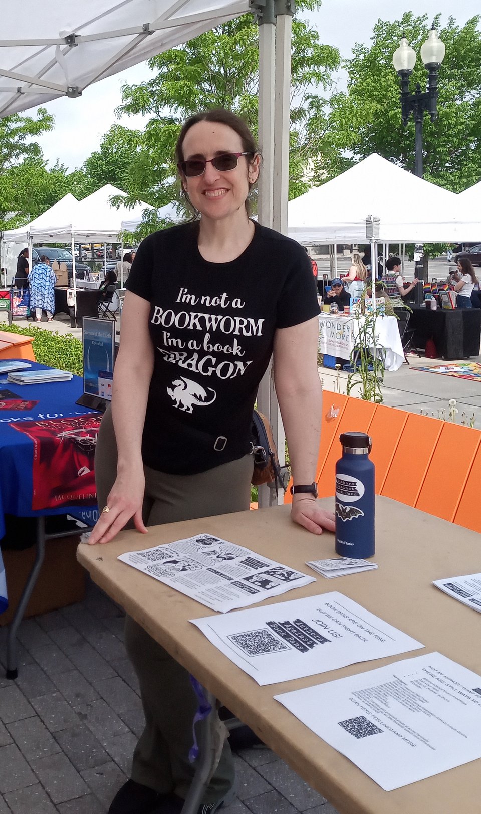 The author, a white woman in sunglasses and a shirt that says "I'm not a bookworm, I'm a book dragon," leaning on a table covered with leaflets.