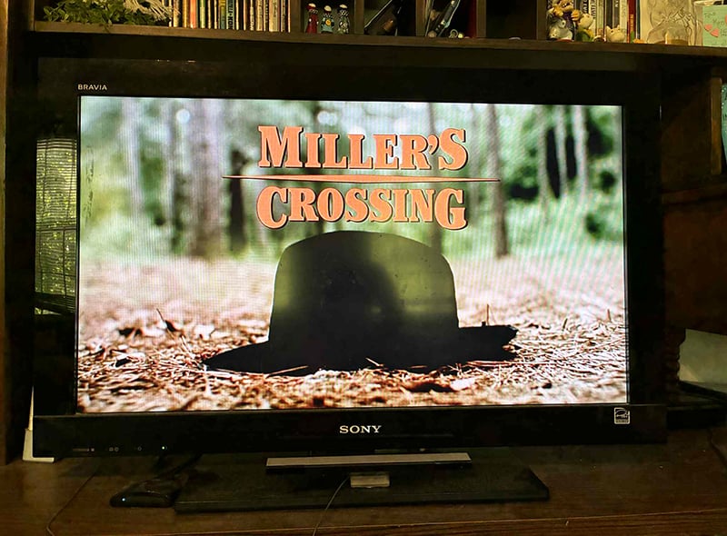 A photo of my television, which in this instant displays the title of the movie Miller's Crossing. There is an image of a hat on the ground in a wooded spot.