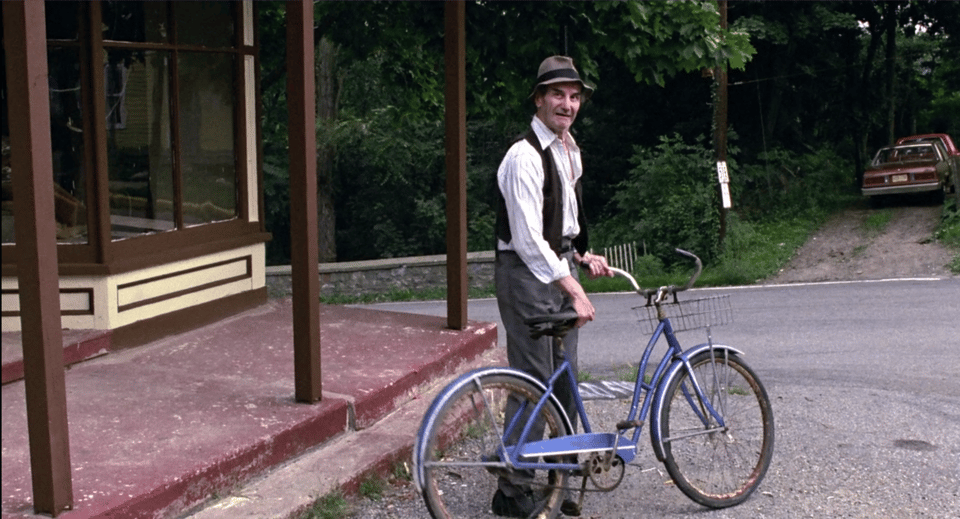 Ralph, a older white man with lank grey hair tucked under a lumpy brown-grey fedora with a rumpled brim and wearing a half-unbuttoned and disheleved white button down with thin dark stripes, a brown vest, rumpled grey slacks, and black shoes looks back at Annie as she leaves the diner. He holds his rusted blue step-through bike with a basket on the front before taking off.