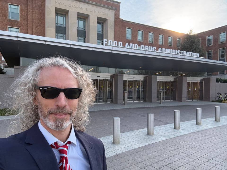 photo of white man in navy suit, light blue dress shirt and red-blue-silver striped tie; He is wearing Wayfarer sunglasses and has wild curly hair. He is standing in front of FDA Building 1.
