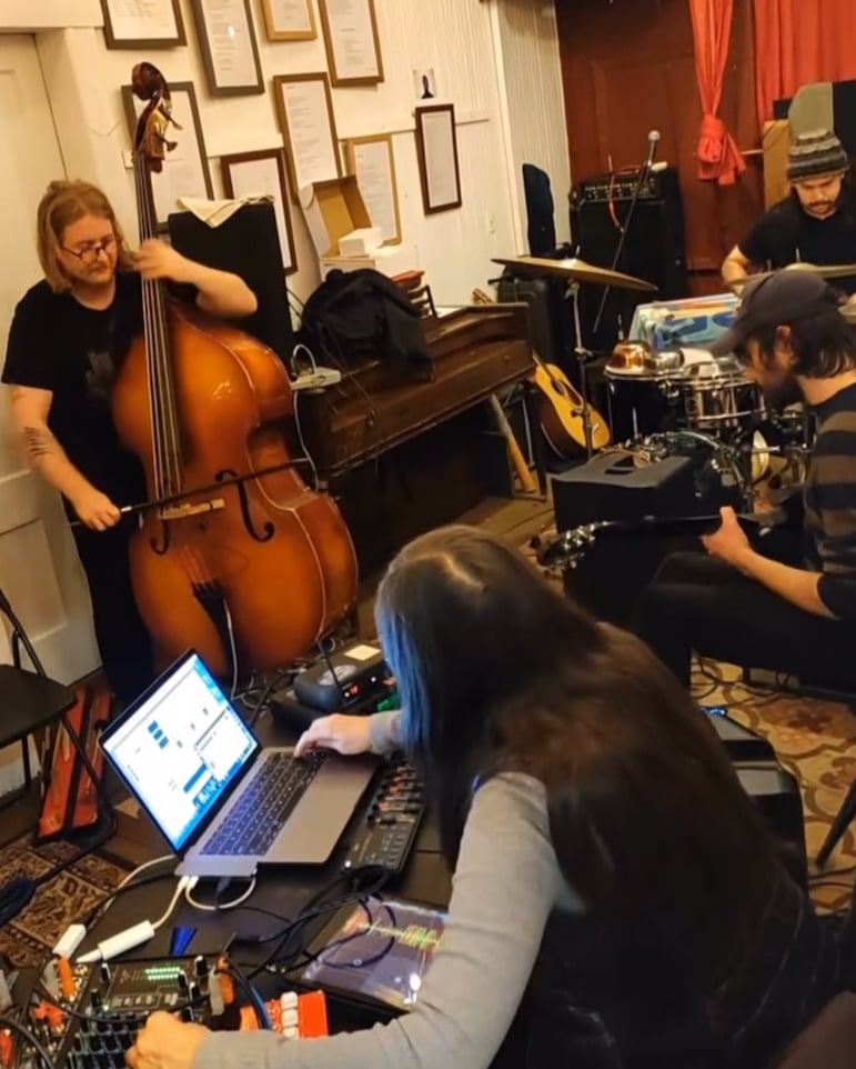 A women in the foreground with her back to the camera presses a key on a laptop computer while a bassist, drummer and guitarist play with her.