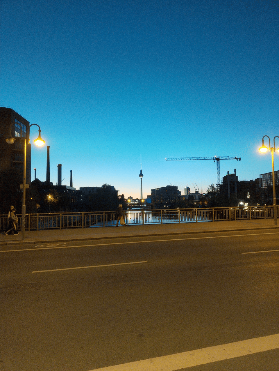 The Berlin TV Tower rises from the center of the background, framed by tall buildings and chimneys on the left and a construction crane on the right. The sky is dark blue at the top, becoming light teal and then peach at the bottom; it's shortly after sunset. The Spree River reflects some of the streetlights. In the foreground is the road. The picture might be better if I'd sprinted across the street, but there's traffic, and I had a train to catch.