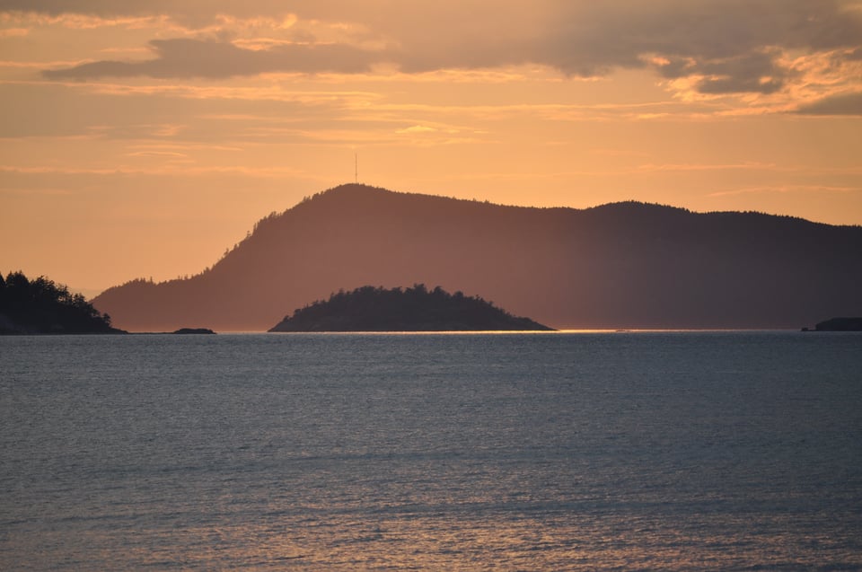 Photo islands at sunset. The foreground is ocean water, with small waves, reflecting some orange light from the sky. The middle of the photo has tree covered islands, some closer than others. The upper third is orange sky with clouds.