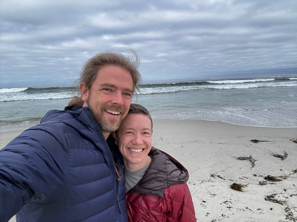 Kal & Drew, smiling, taking a selfie on a windswept beach, bundled in puffer jackets against an overcast sky, gentle waves rolling onto pale sand scattered with seaweed