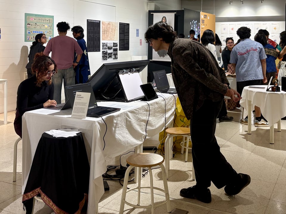 An interactive installation comprising a set of individual laptop computers, an iPad, and a television screen laid out on a rectangular table with a white cloth. Two people are engaging with the exhibit which also has a stool covered in a black saree, constituting its own surface for handouts. The installation includes stools for people to sit and interact with the objects.