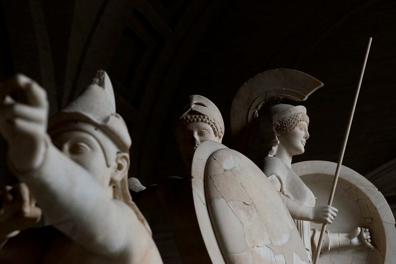 A photo of several ancient Greek sculptures of Warriors. One appears to be Athena - on the right hand side, facing towards the right. The other two are men, one holding a shield and one raising a bow and arrow, and they are facing towards us. The background is very dark black, and they are lit from the right hand side, so they stand out in sharp contrast against the background. It is a very dramatic effect.