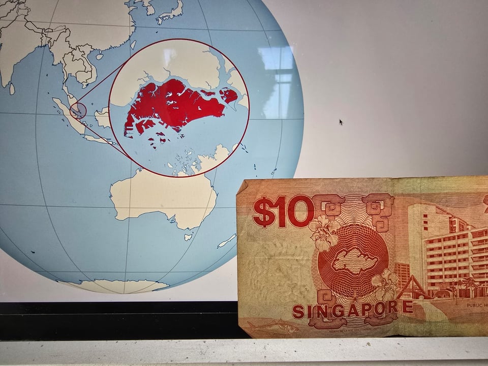 A 10 dollar note from Singapore placed on a laptop screen. The note shows a picture of a map of Singapore in red. The screen displays a map of South-east Asia, with a zoomed-in circle showing Singapore highlighted in red. The map on the screen is noticeably different to the one on the note.