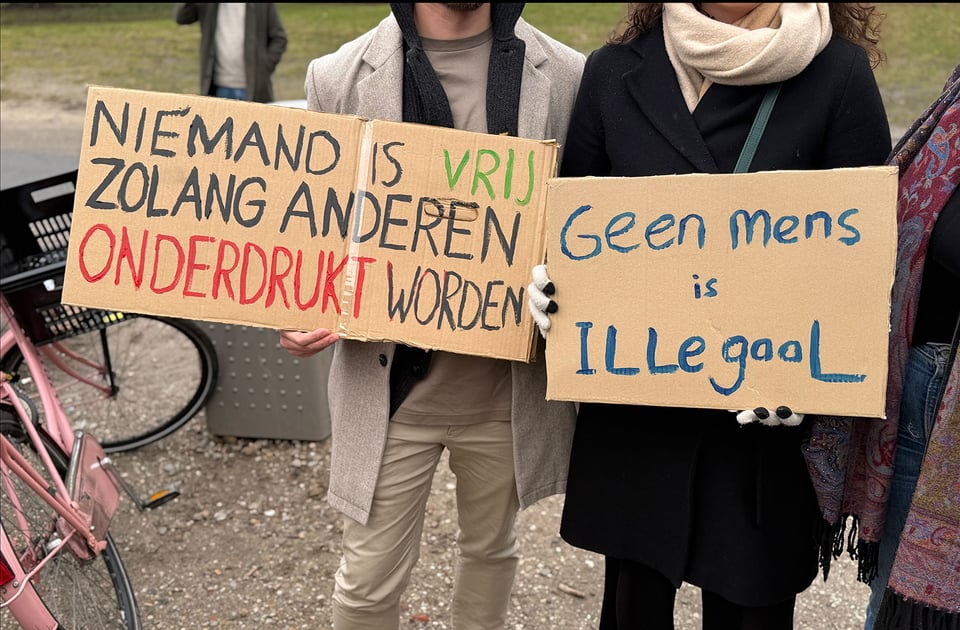 Two people hold up signs in Dutch:
1 - Niemand is vrij zolang anderen onderdrukt worden
2 - Geen mens is Illegaal