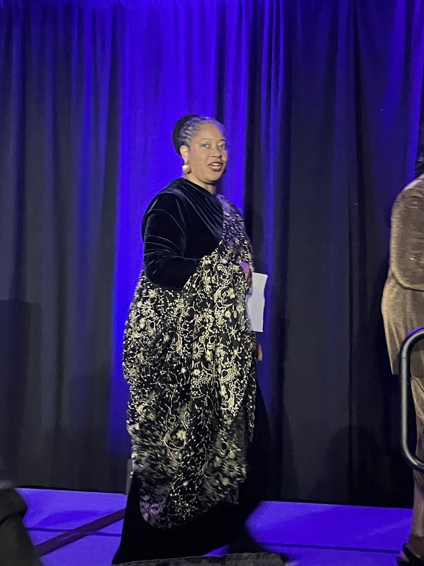 At the 2026 Nebula Awards in Rosemont, Illinois, N.K. Jemisin the Grandmaster walks to the stage during the awards ceremony. She has her hair in a bun with bronze pendant earrings and a black dress embroidered with gold constellations.