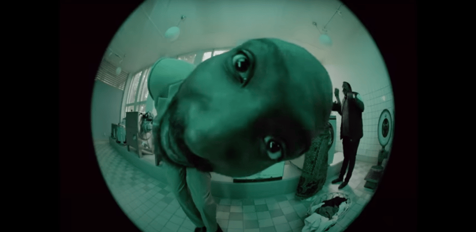 A distorted, close-up photograph taken with a fish-eye lens, showing a person wearing a full green suit and whose face is very close up to the lens. They are standing in a room that appears to be a medical or laboratory setting, with tiled walls, medical machinery, and a partially visible person in a dark suit in the background.