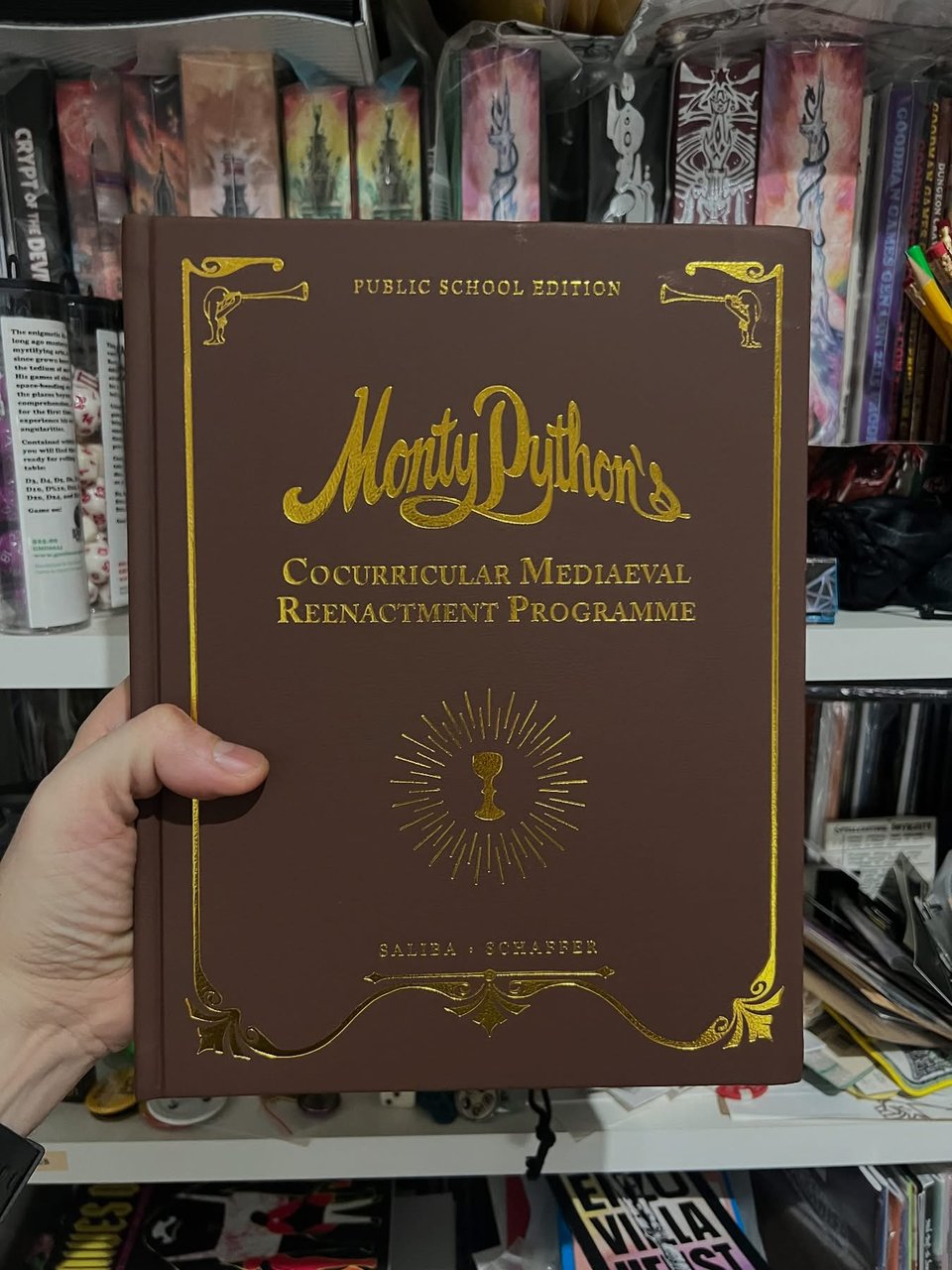 A hand holding a leatherbound book, with gold details where you can read the title of the book and see the holy grail