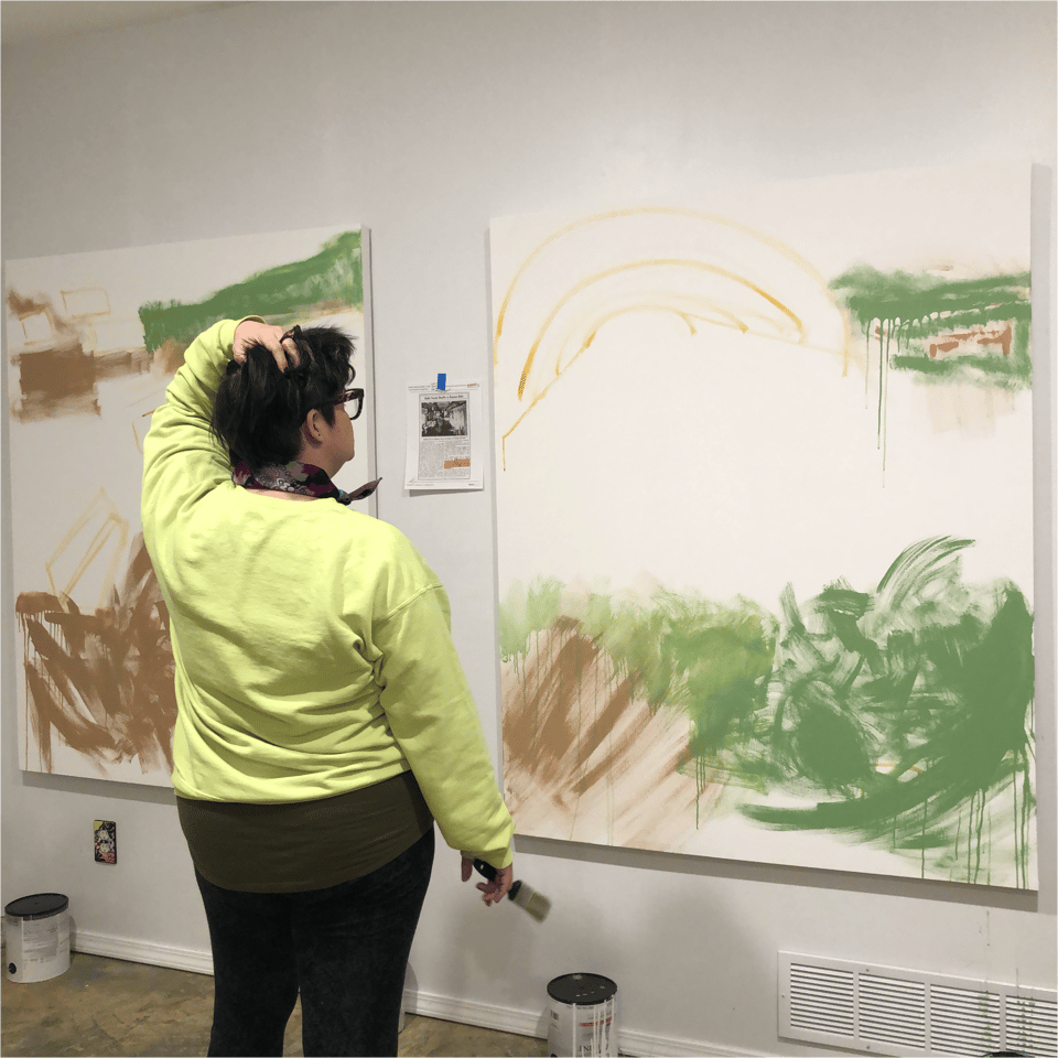 the artist KCF stands in front of two canvases with messy brush strokes on them and is thinking about their next move