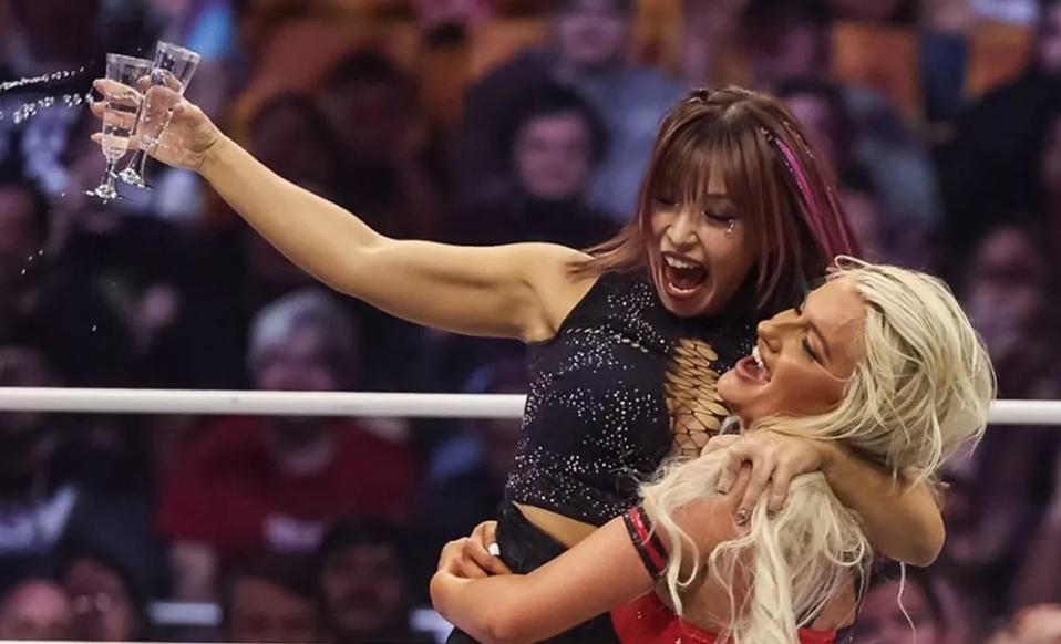 Mariah May holding up Mina as she spills champagne from two glasses in her hand.