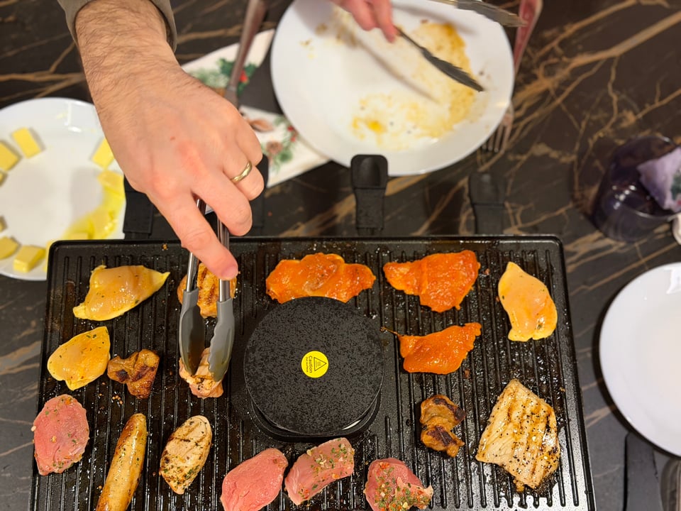 A hand uses tongs to turn over a small piece of meat on a grill on a dining table covered in over a dozen small pieces of meat.