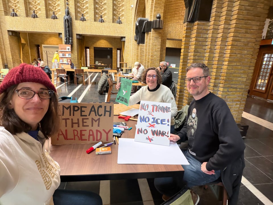 Three people sit around a table with hand made signs they made together. "Impeach them already" "No Tyrants X No ICE! X No WAR!"