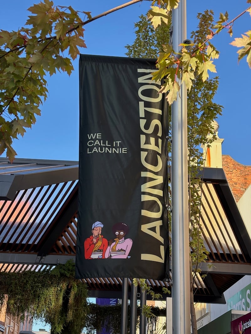 A little sunny street, lots of plants, and in the foreground a big banner reading LAUNCESTON and WE CALL IT LAUNNIE, with a little cartoon drawing of two people eating noodles