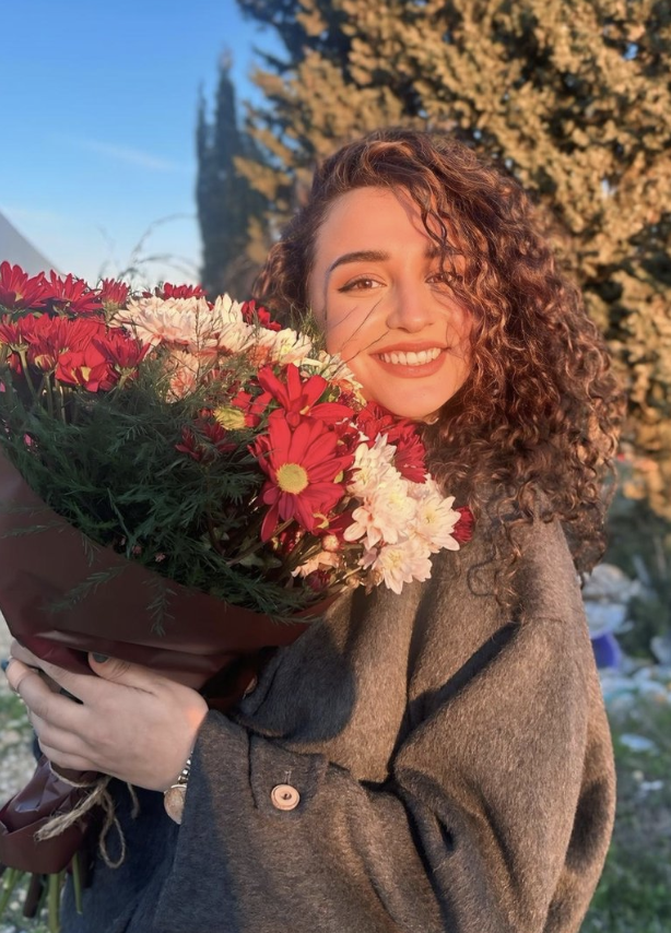 A beautiful young woman with long brown curly hair and a big smile, holding a bouquet of flowers