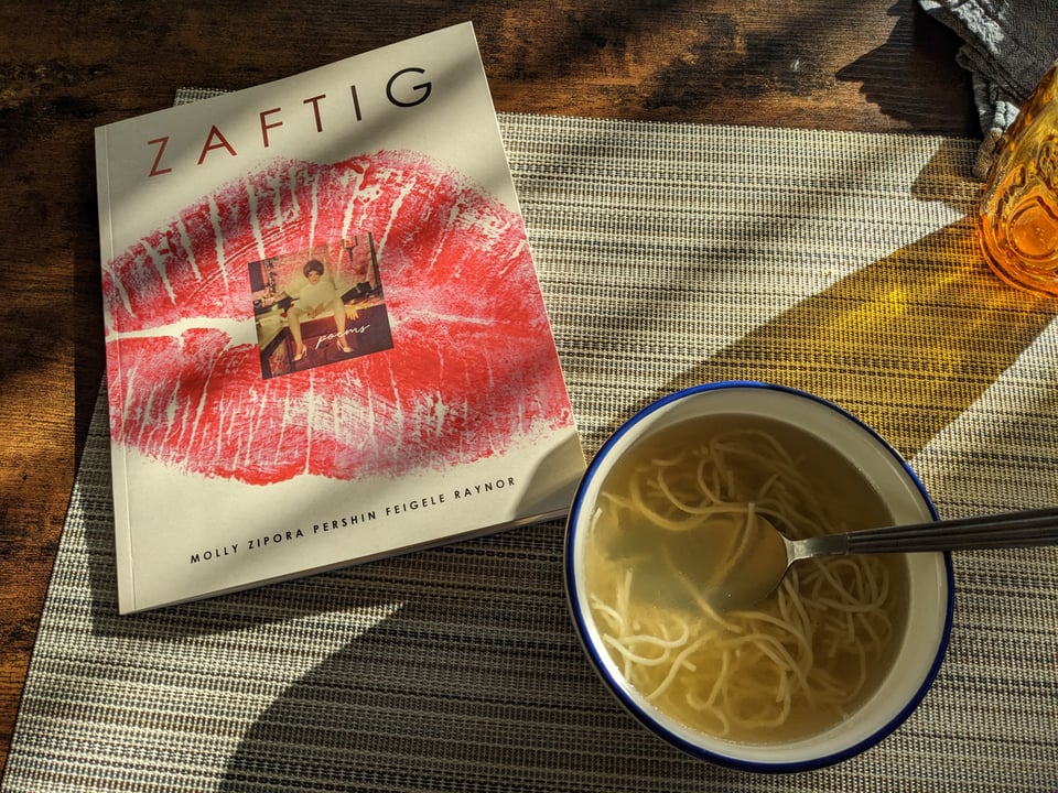 Zaftig by Molly Raynor, next to a bowl of chicken noodle soup. The book cover is a bright pink lipstick kiss that takes up the entire cover, overlaid with a color portrait of Raynor's grandmother.
