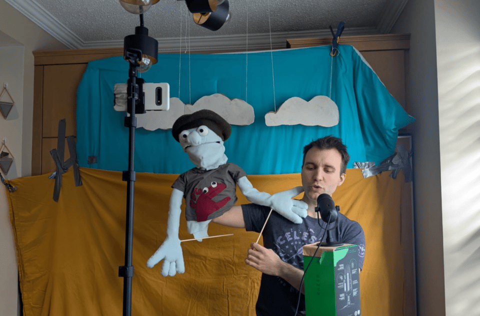 Kurt stands in front of a backdrop, puppeteering Herman. A mic is set up in front of him on top of a box, and behind him there's sheets duct-taped to the wall. Herman is awkwardly angled, stroking Kurt's face.
