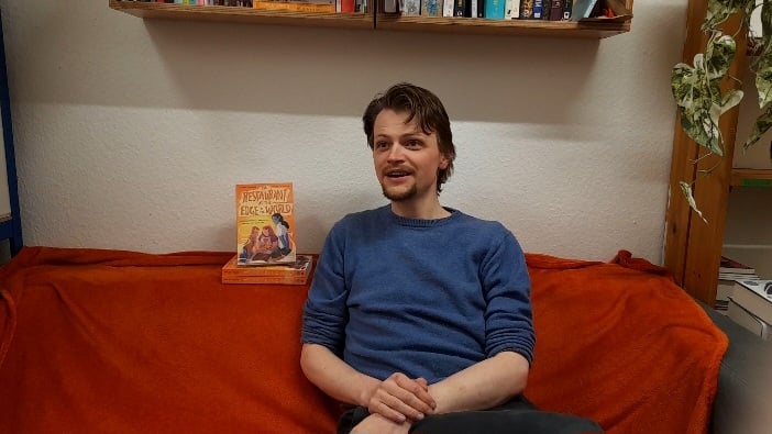 Me, sitting on an orange sofa, talking to someone off camera. Two copies of THE RESTAURANT AT THE EDGE OF THE WORLD are next to me.