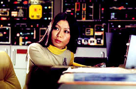 A Japanese woman with shoulder length black hair, wears a tan jump suit with a yellow collar, sits behind a desk, in the background is a wall of technical readouts, displays and controls.