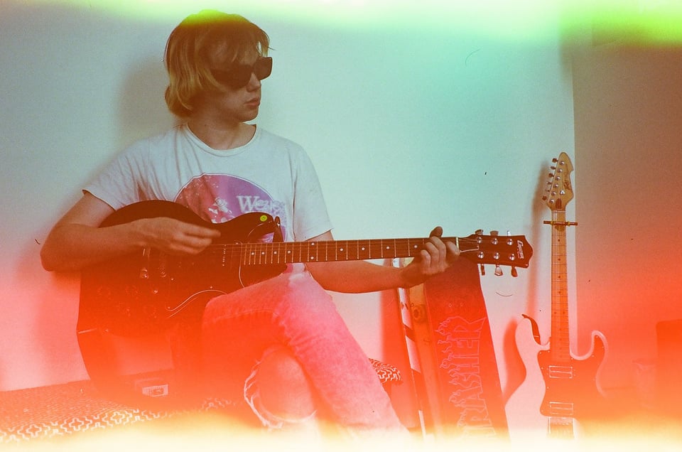 A young wearing sunglasses sits with his right leg crossed over his left, playing an electric guitar.