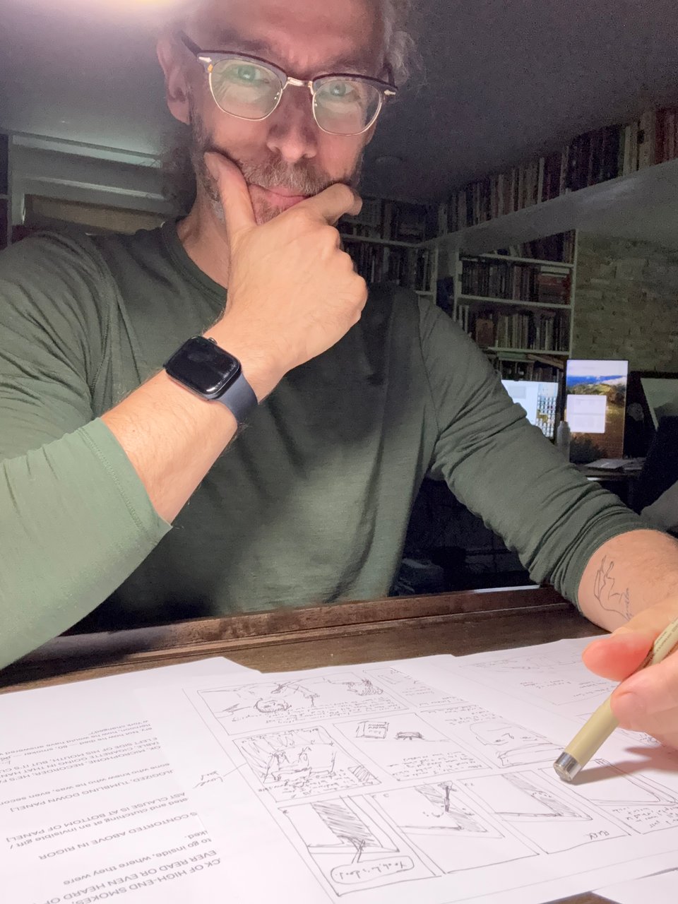 photo of white man in glasses & a green pullover at a drawing table, tapping a micron pen on some comics layout pages