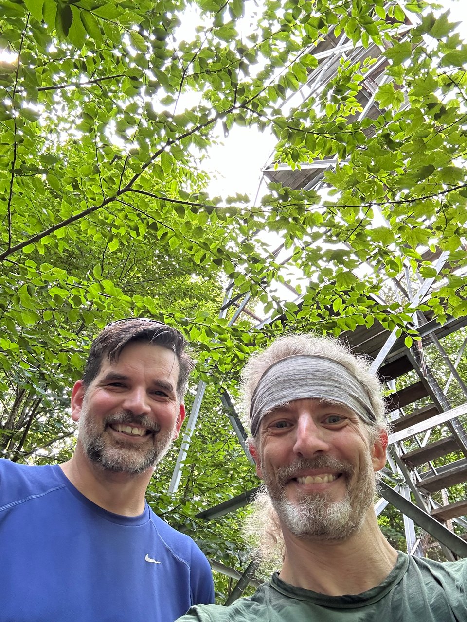 Two white men in the foreground, with trees and green leaves behind them and a metal fire tower looming above them