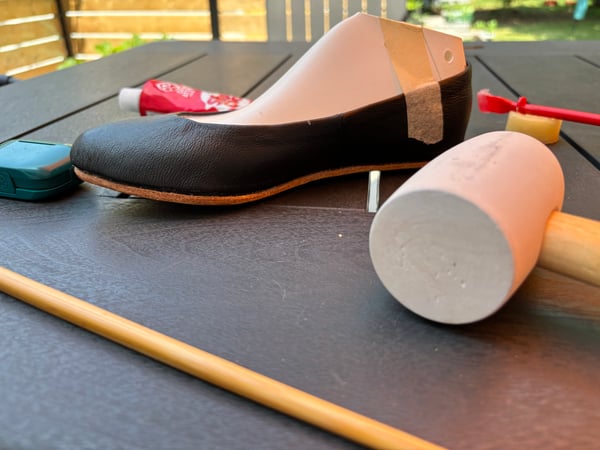 A nearly finished shoe in the process of being lasted, with a black leather upper and natural veg-tanned leather soles, surrounded by an assortment of tools