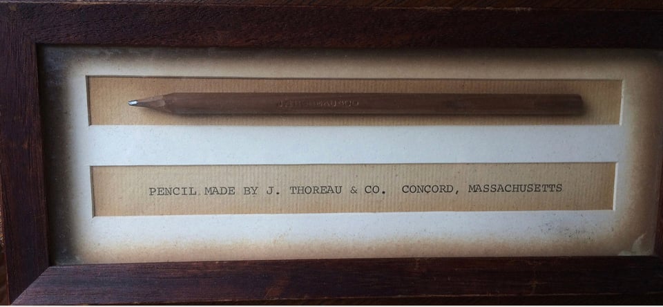 A close up photo of a museum case holding a very old thick wooden pencil, with a crudely sharpened point. The text mounted below the pencil reads "pencil made by j. thoreau and co., concord, masachusetts"