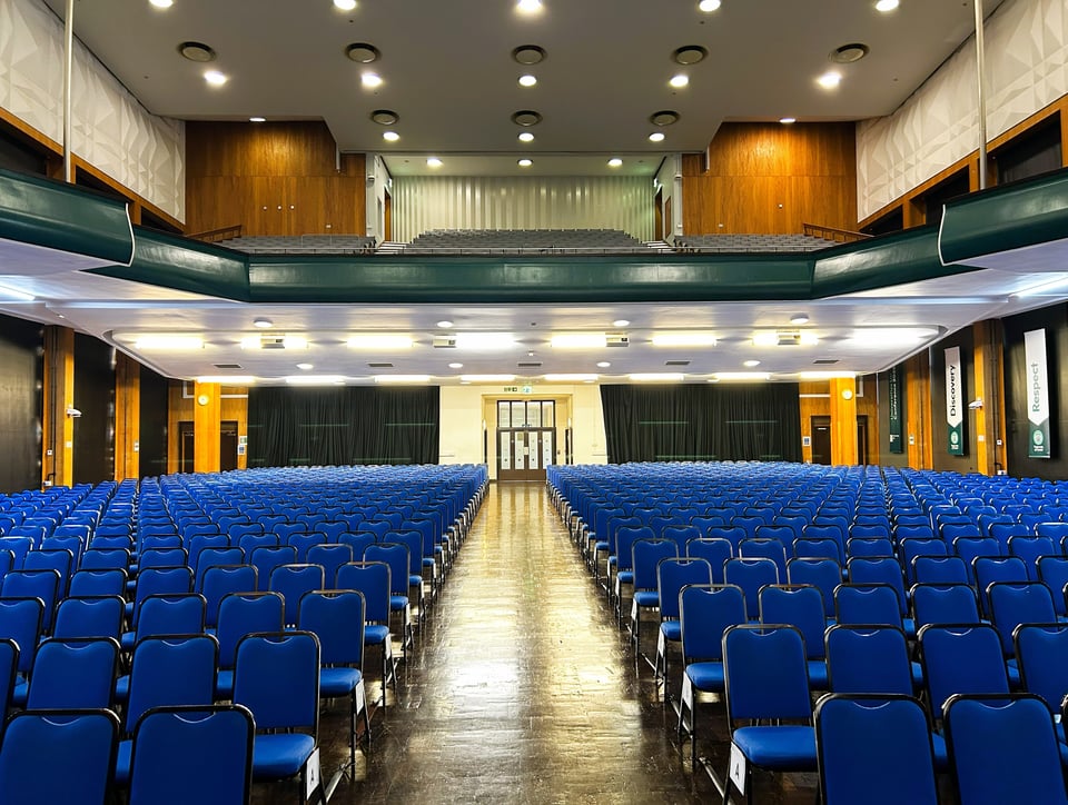 The interior of the Great Hall at Uni of Exeter, laid out with rows of chairs for the graduation. It's a 1960s hall with a single circle and lots of Norwegian redwood or similar. The architecture is symmetrical. The aisle in the middle of the chairs is slightly to the left, meaning the whole photo looks asymmetrical.