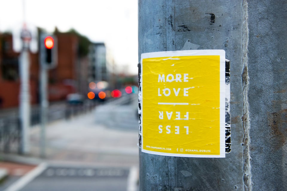 A photo where on the right side in the foreground is a light poll with a yellow sticker with white writing. The sticker says "More love" and then a white horizontal line and then "Less Fear" written upside down and backwards below. It gives the visual appearance of the mirror version of more love being less fear. To the left is the out of focus view down the street past the pole, including several traffic lights fading into the distance.