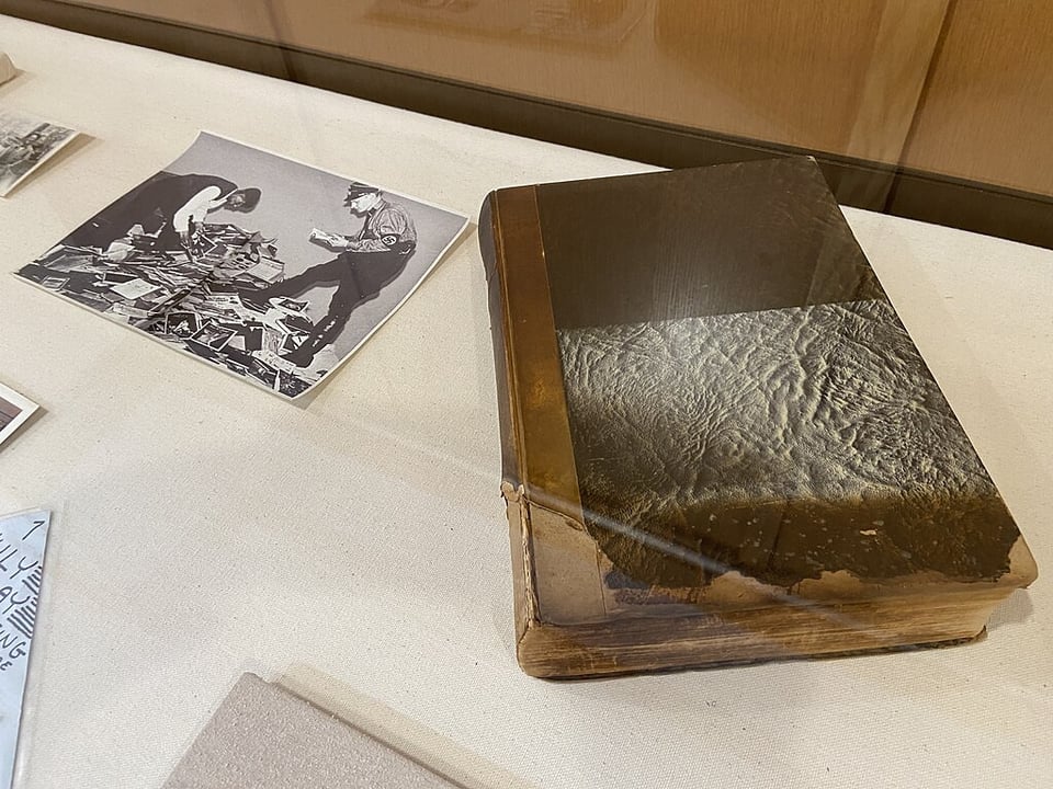 A dark brown book, its pages curled slightly by fire and damaged by smoke. It's under a glass case, alongside a photo of two men, one with a Nazi SS armband, crouched over a pile of documents and books from the Institute. Attribution: By Myotus - Own work, CC BY-SA 4.0, https://commons.wikimedia.org/w/index.php?curid=133507974