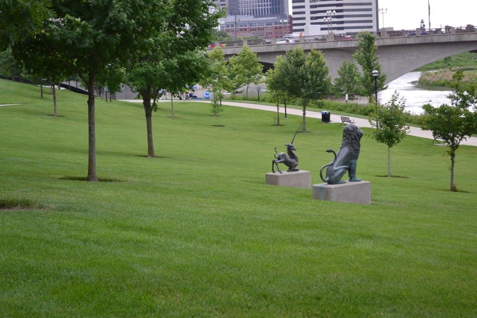 A large lawn by a river. Two black statues of a seated lion and a bowing unicorn face the river. In the distance is a concrete bridge and some high rise buildings.