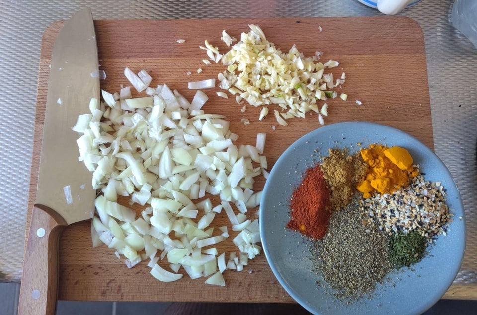 Image description: top-down photo of a cutting board, with a knife on the left, piles roughly minced garlic and onion in the middle, and a small blue plate filled with colorful piles of spices on the right. end image description.