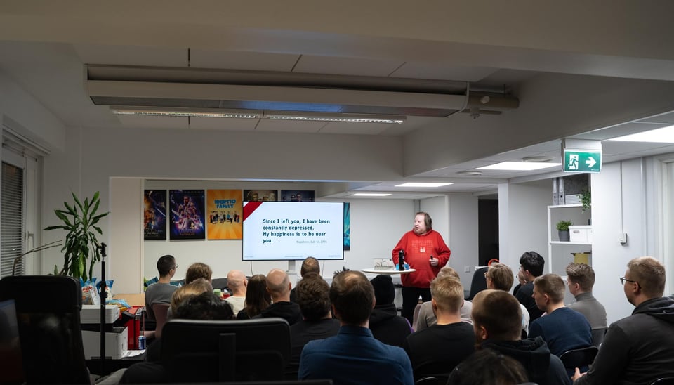 Juhis speaking to a room full of people, wearing a bright red hoodie and on the slide there's a quote from Napoleon's love letter: Since I left you, I have been constantly depressed. My happiness is to be near you.