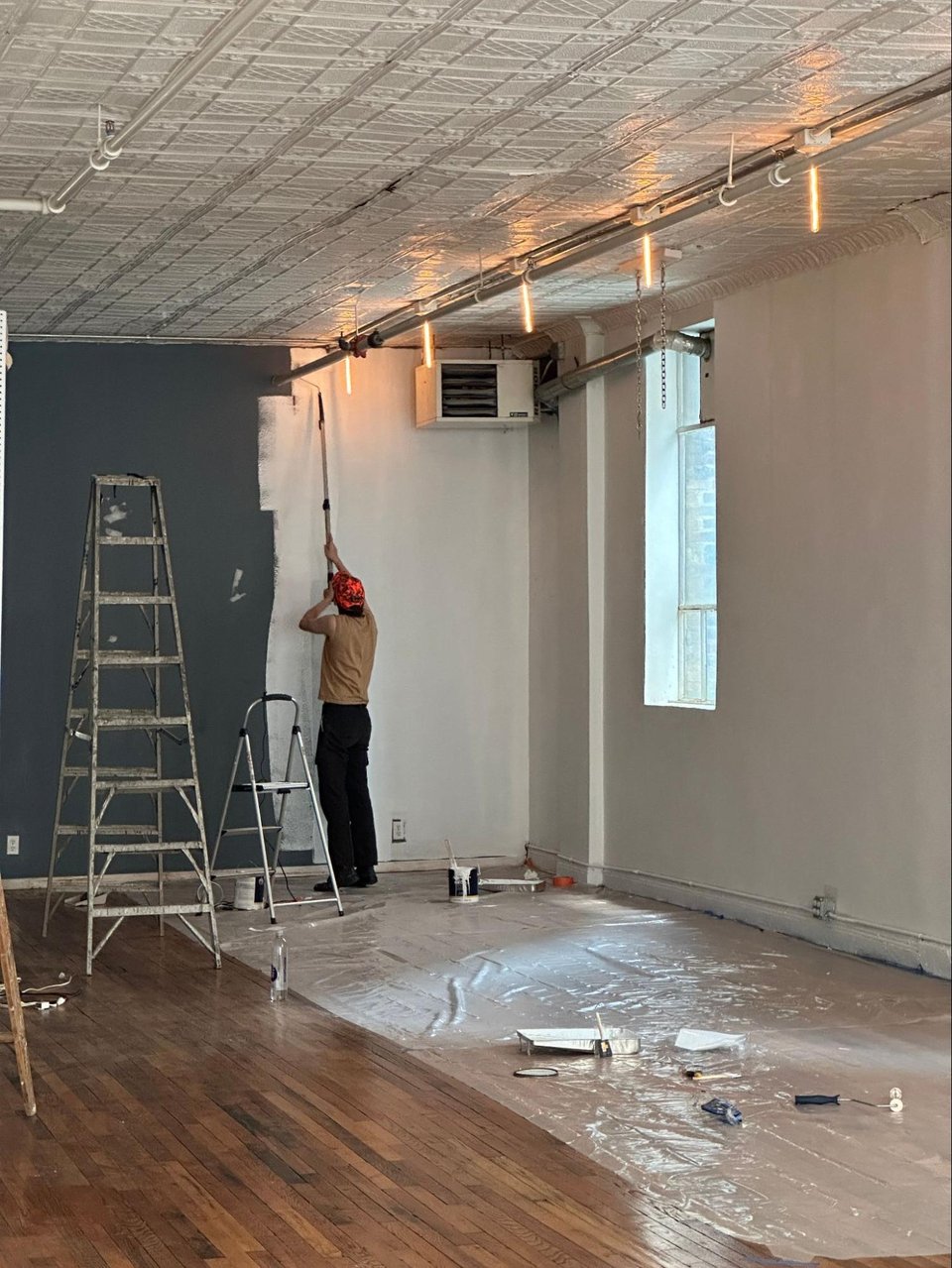 A person painting the space white. A ladder, paint cloth, and rollers covering the floor.