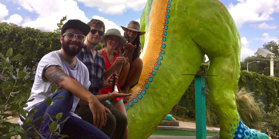 Four improvisers sit on the tail of a T-Rex model at a mini-golf tournament.