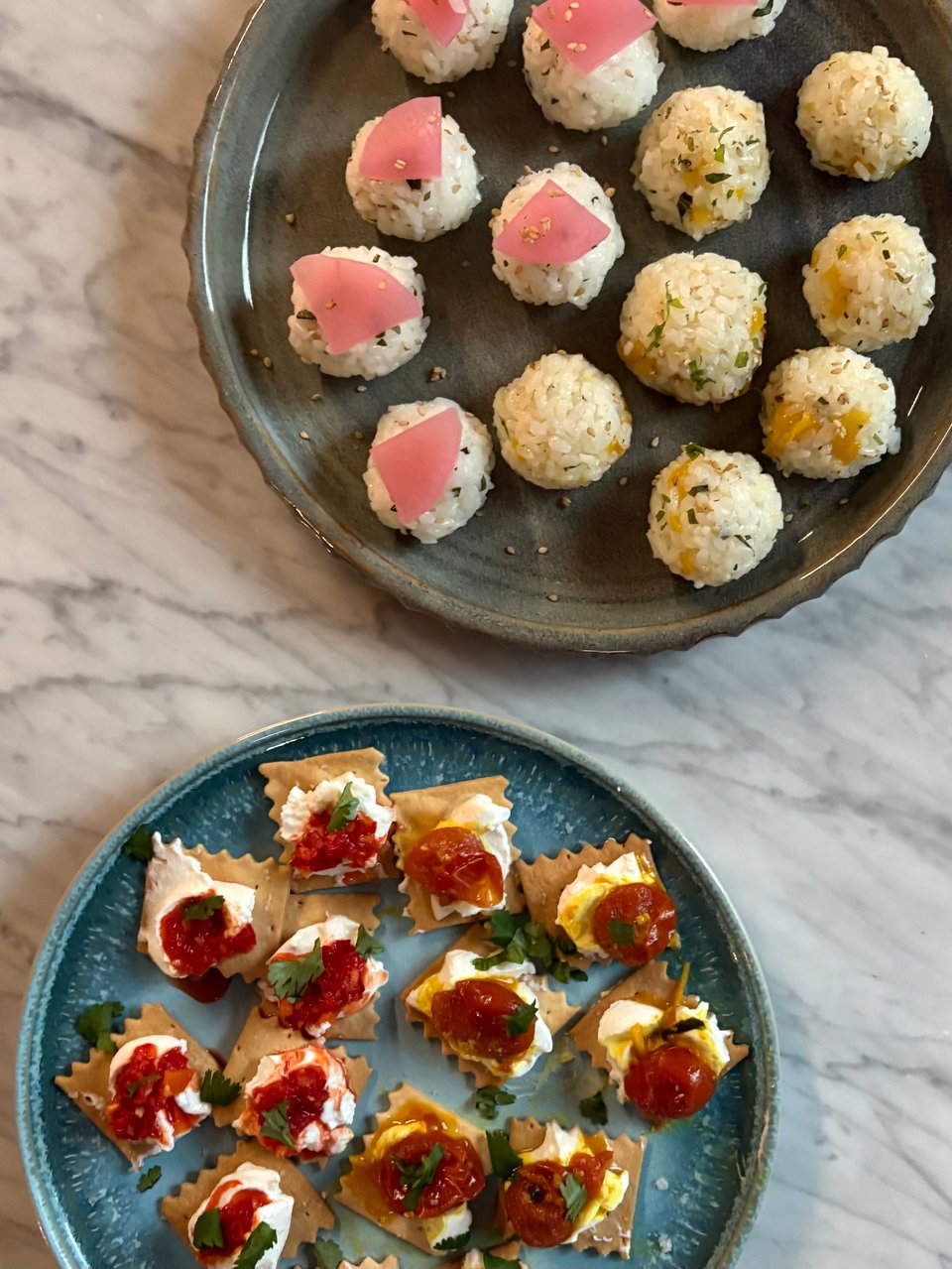 Two kinds of onigiri (mango jam, shiso, & sesame and shiso, sesame, & Daikon pickle) on a blue-gray platter and two kinds of cracker snacks (labneh topped with red chile condiment & labneh topped with ginger-garlic cherry tomatoes) on a blue plate.
