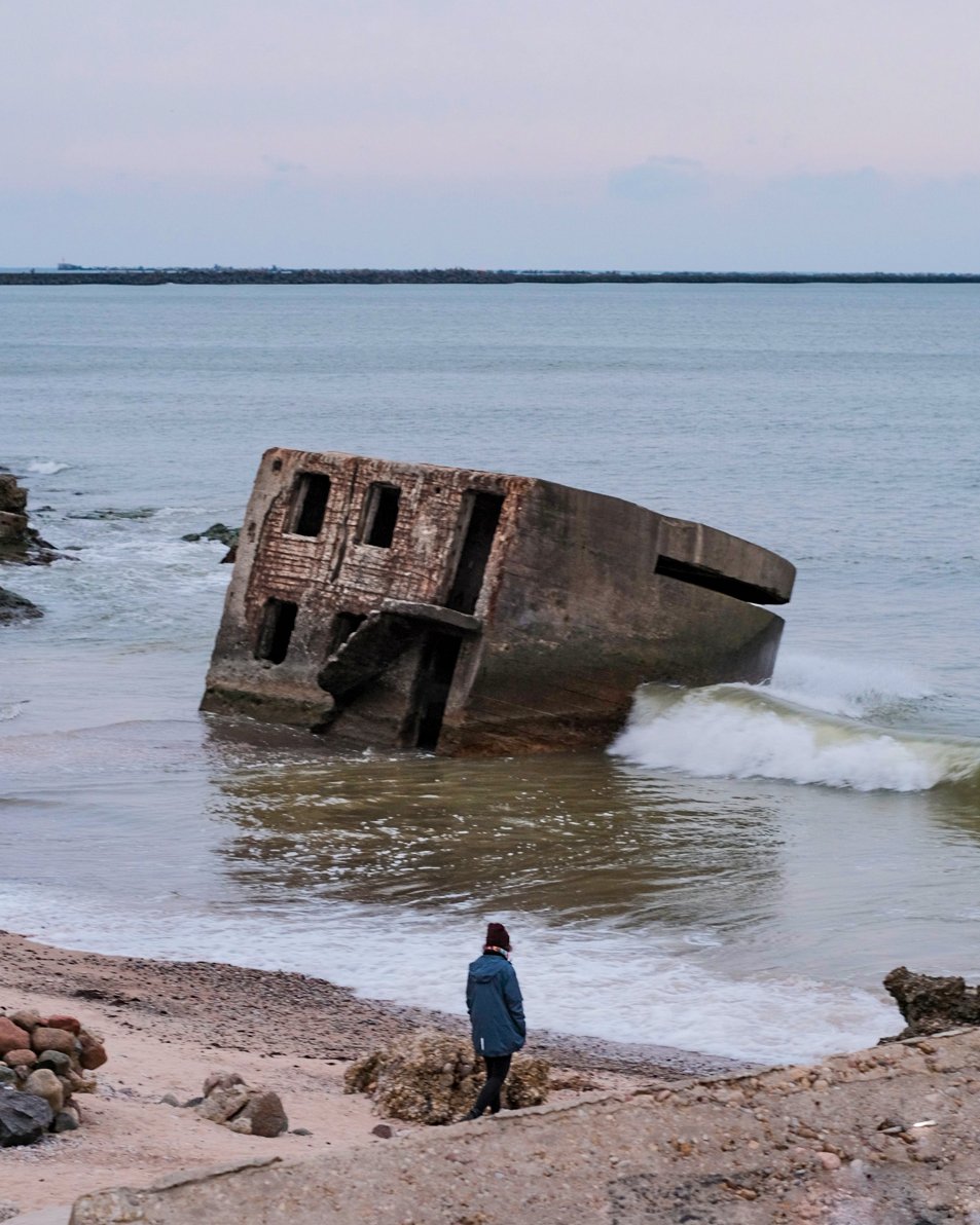 A photo of a person standing on the beach. Beyond them in the water, waves crash against a half-submerged concrete building.