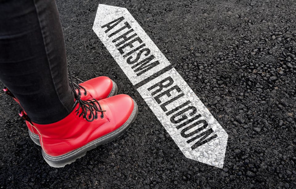Imagine of a feet wearing red boots upon tarmac. Two arrow signs are painted on the road, facing in opposite directions. One reads "atheism", the other reads "religion".